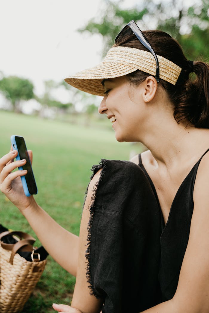 A woman in a sun hat smiles during a video call, seated on grass in a park.